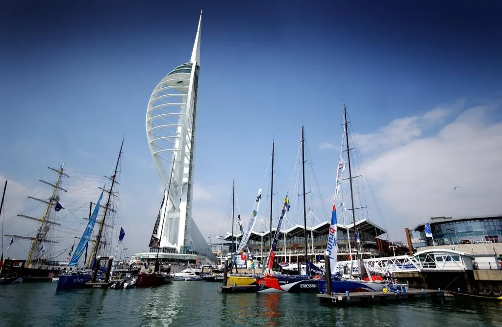 This is a photograph, not a designed graphic.

Tall ships and modern sail boats moored in a harbour with a distinctive white curved tower building in the background under blue sky.