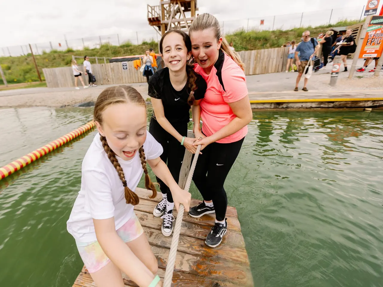 Three young people on a wooden platform by water, holding a rope during an outdoor activity session.