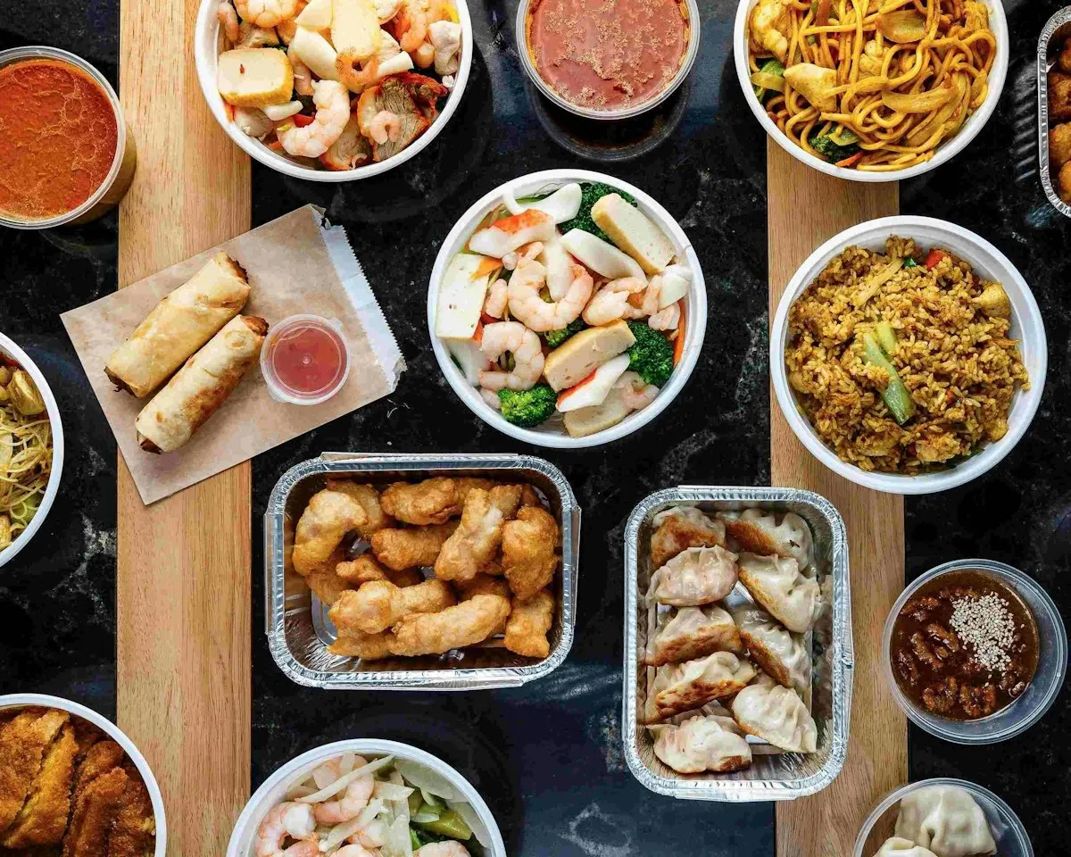 Overhead view of various Asian takeaway dishes including spring rolls, fried rice, noodles, dumplings, and seafood with sauce bowls.