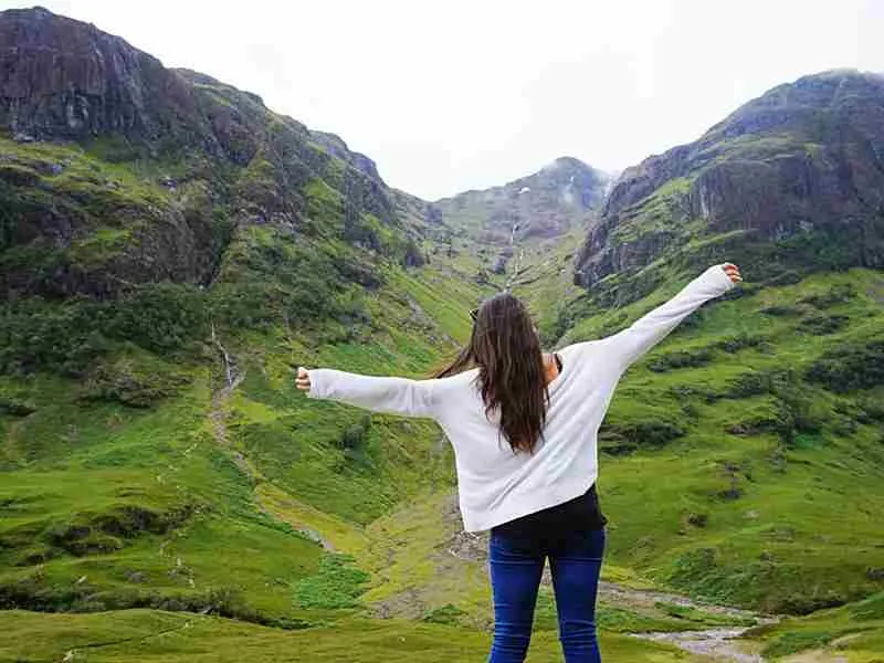Woman standing with arms outstretched in a verdant Highland valley with rocky slopes on either side.
