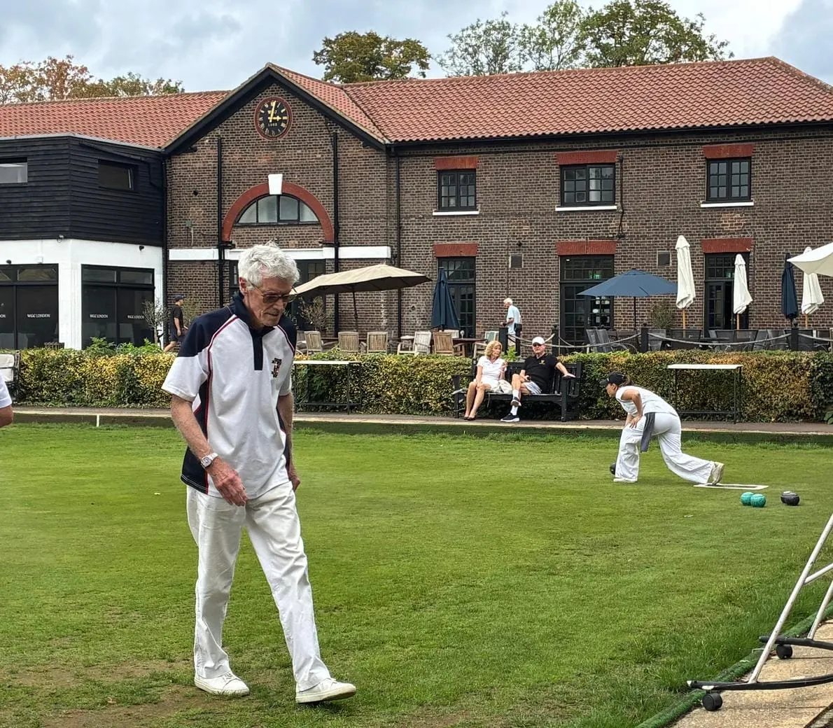 Older man in white clothing playing lawn bowls on a green in front of a brick clubhouse, with other players and spectators nearby.