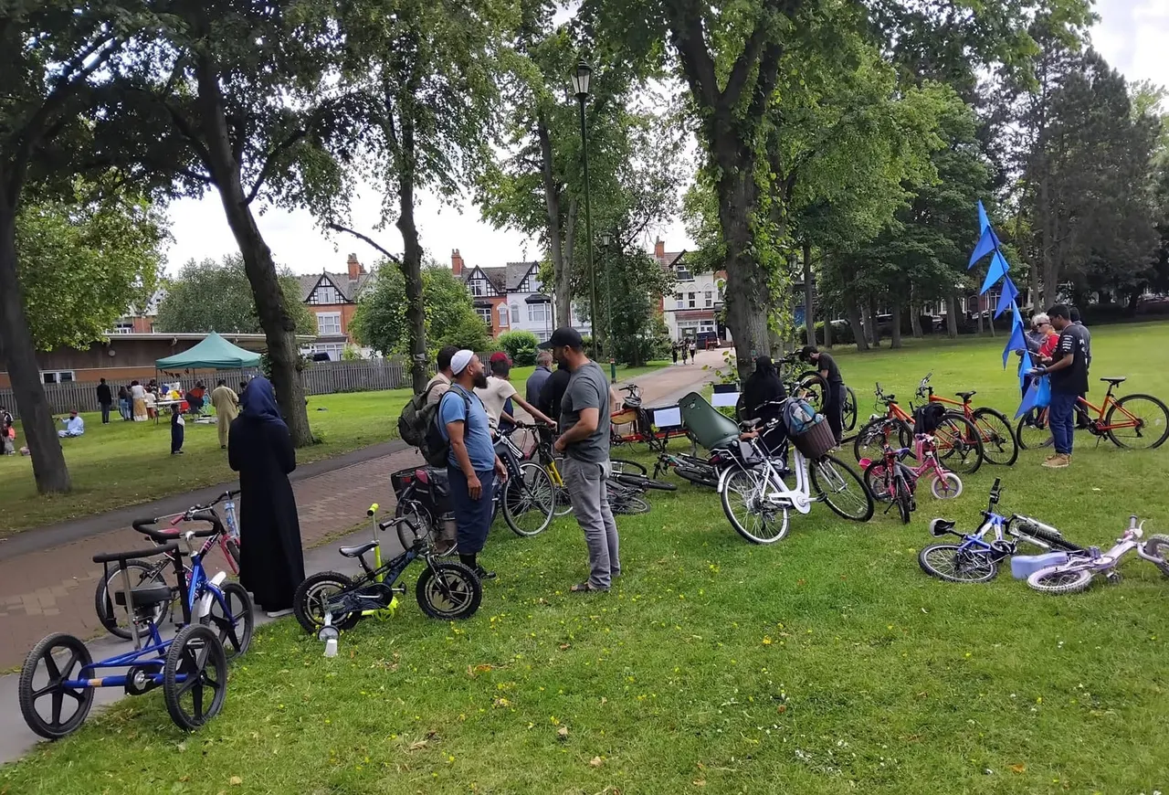 A community cycling event in a park with children and adults gathered around bicycles of various sizes on a grassy area beneath large trees.