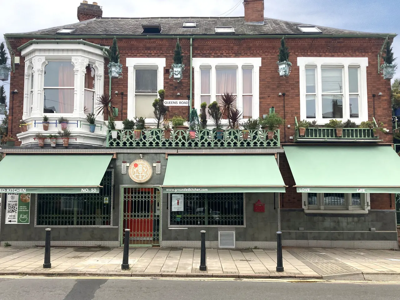 Victorian brick building with ground floor restaurant and mint-green awning, ornate bay window and balcony with potted plants above.
