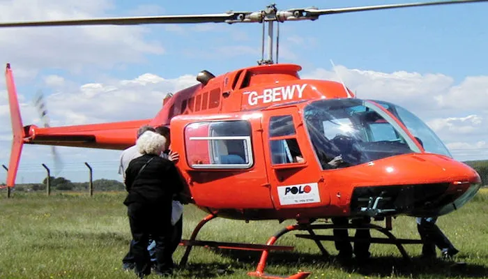 An older person in dark clothing approaches a red helicopter parked on grass.