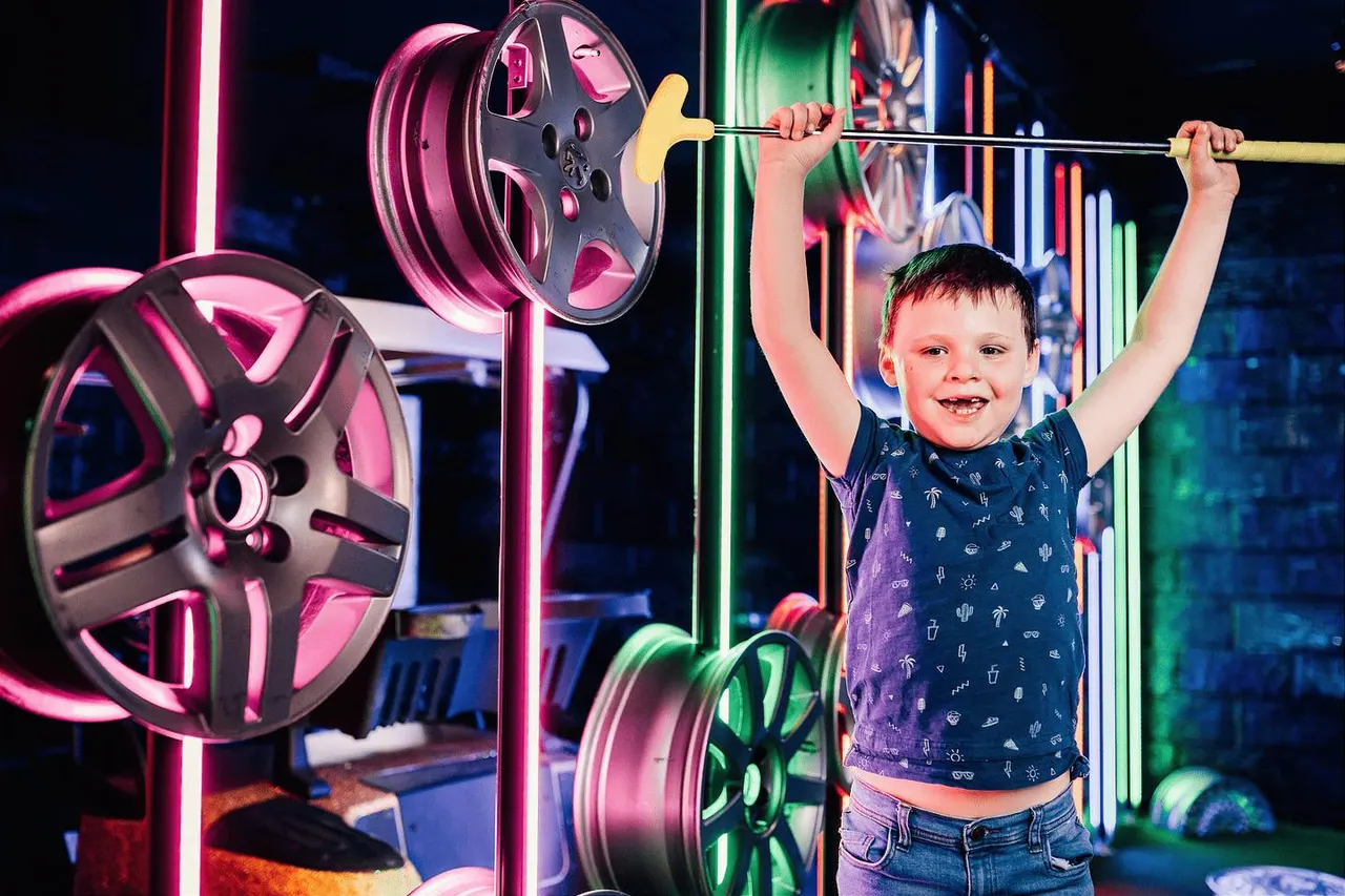Young child holding a weightlifting bar with neon-lit weights in an arcade or entertainment venue.