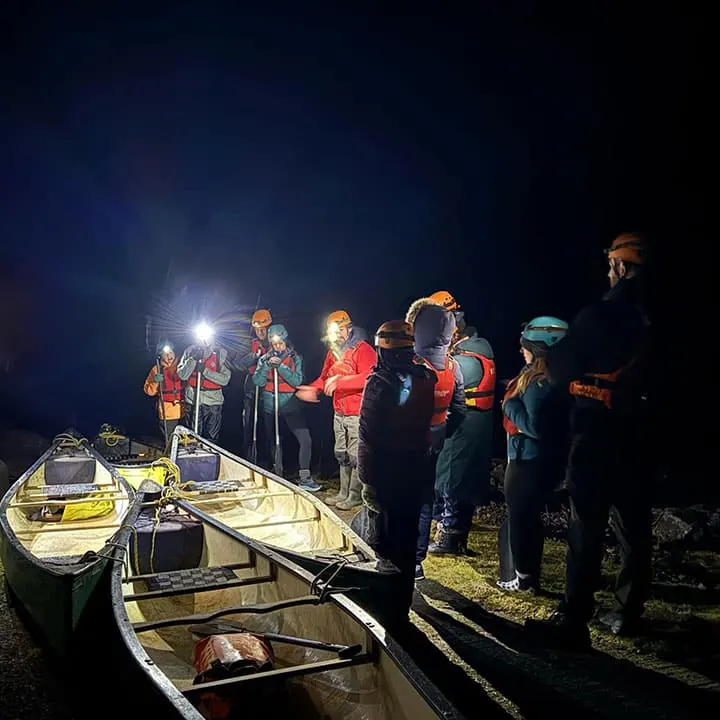 A group in safety gear and life jackets gathered at night beside boats on a river, illuminated by floodlights during a caving or gorge activity.