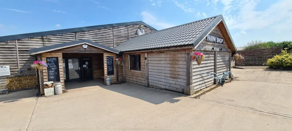 Rustic farm shop buildings with weathered wooden cladding, signage, and hanging baskets on a sunny day.