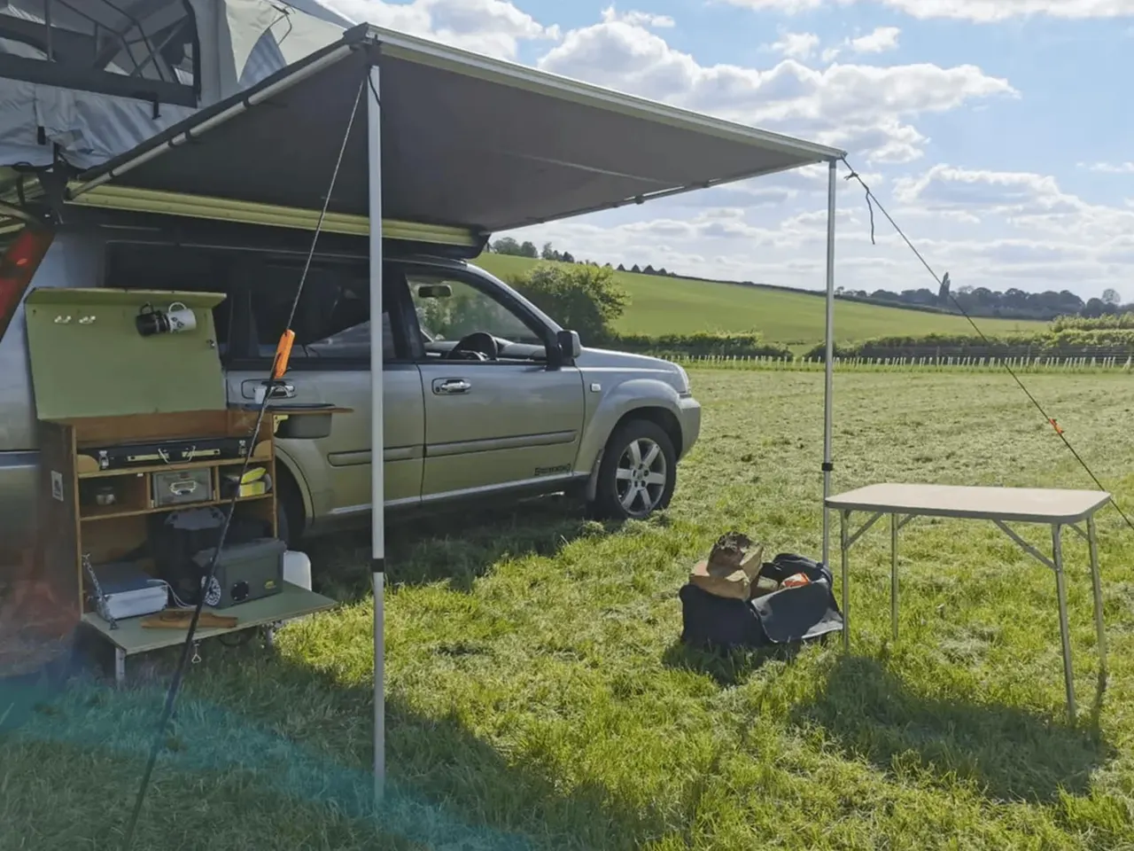 Camper van with awning set up in a rural grassland field, with a portable kitchen and table for outdoor camping.
