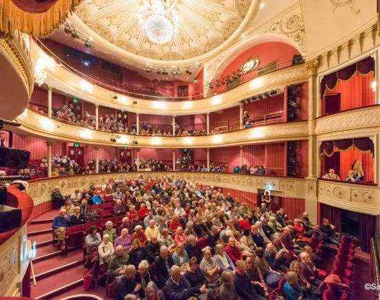 A theatre auditorium filled with seated audience members facing the stage, with ornate balconies and gilded interior.