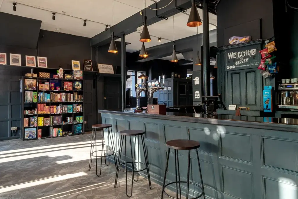 Modern games café interior with service counter, bar seating, board game shelves, and pendant lighting.