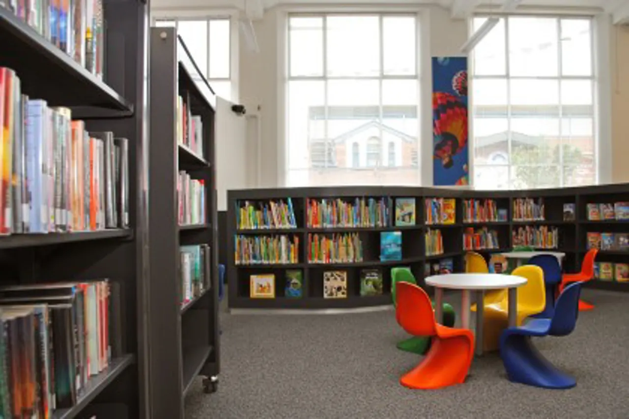 A bright library interior with black bookshelves, colourful modern chairs, a round table, and large windows overlooking buildings outside.