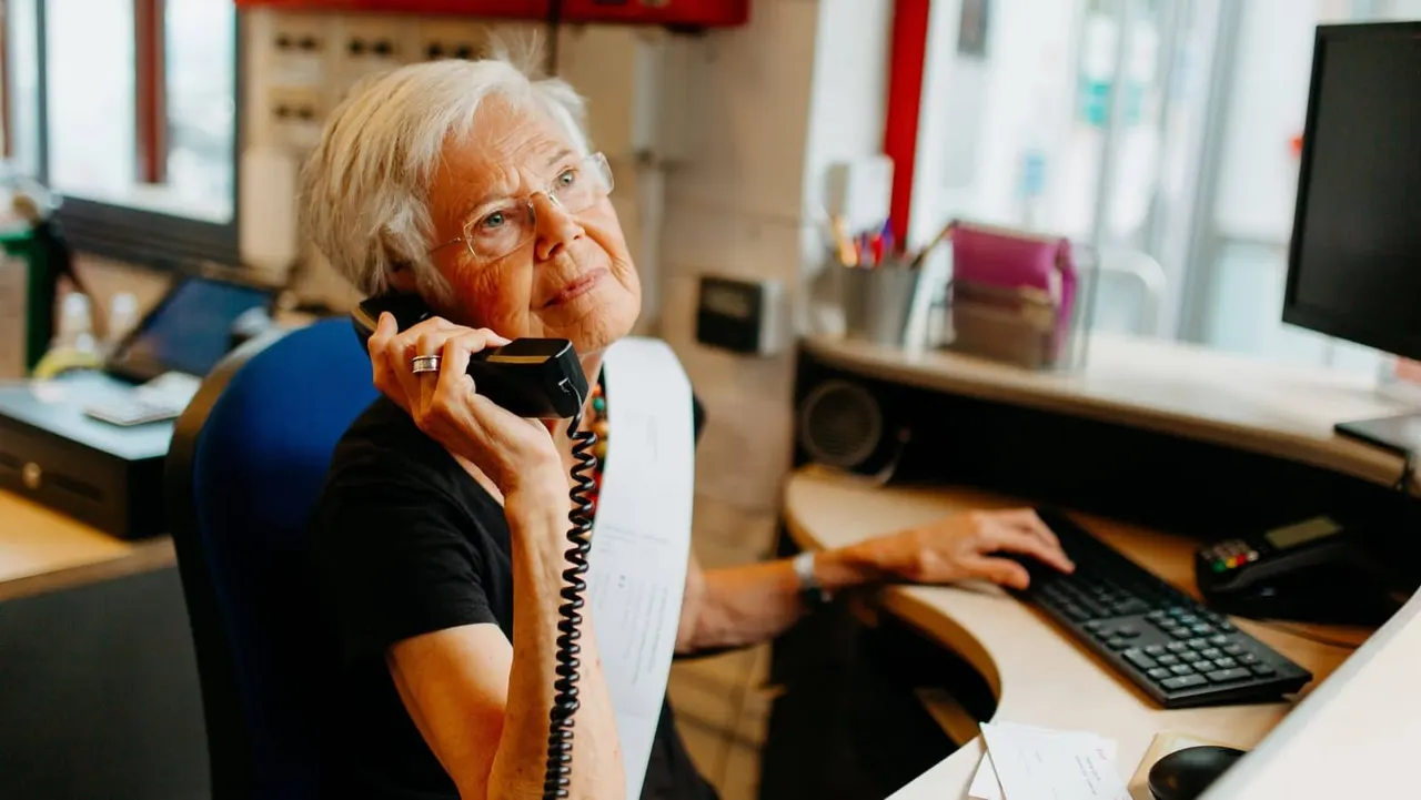 Older woman with white hair and glasses sitting at a desk, speaking on a telephone handset whilst using a computer keyboard.