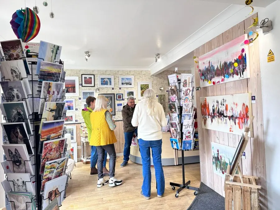 Visitors browse artwork displayed on walls and rotating stands in a bright gallery space with wooden flooring.