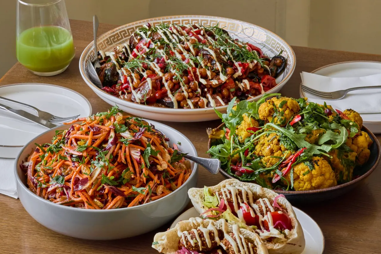 A table set with multiple colourful sharing dishes including roasted vegetables, wraps with drizzled sauce, and a green juice drink.