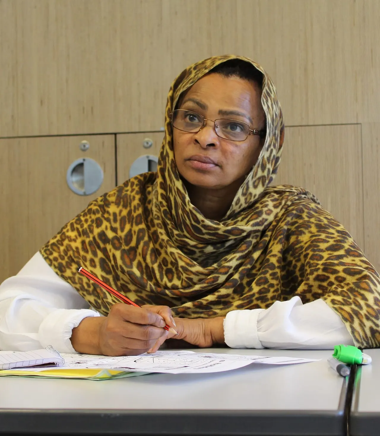 Woman wearing glasses and a patterned headscarf sitting at a desk with papers and a pen.