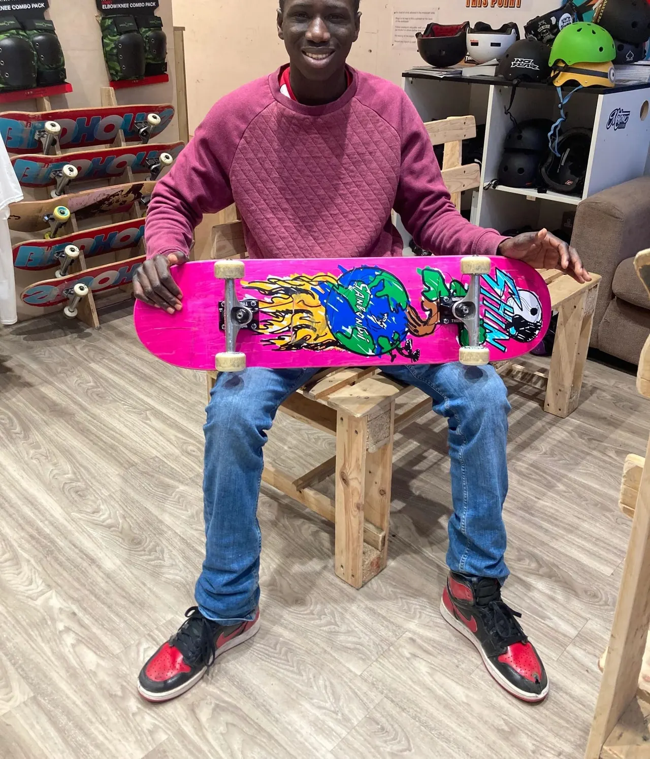 Man holding a bright pink skateboard with colourful graphics in a skate shop surrounded by equipment and other boards.