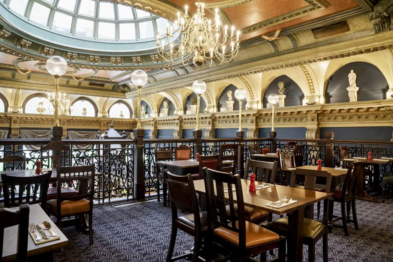 Ornate Victorian dining room with domed glass ceiling, crystal chandeliers, decorative busts, and wooden tables set for dining.