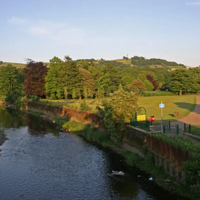 River running alongside grassy fields with tree-covered hills beyond and station infrastructure visible on the near bank.