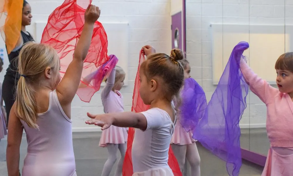 Children in pink dance costumes playing with red and purple scarves during a dance class.