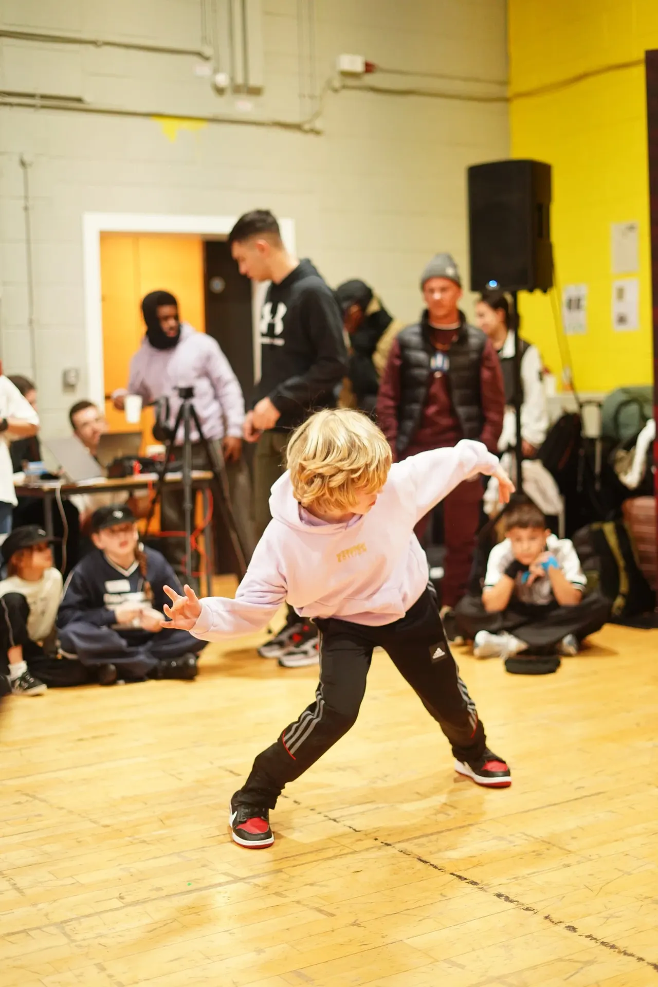 Young child performing a dance move in the centre of a wooden floor whilst seated spectators and standing adults watch in a bright studio.