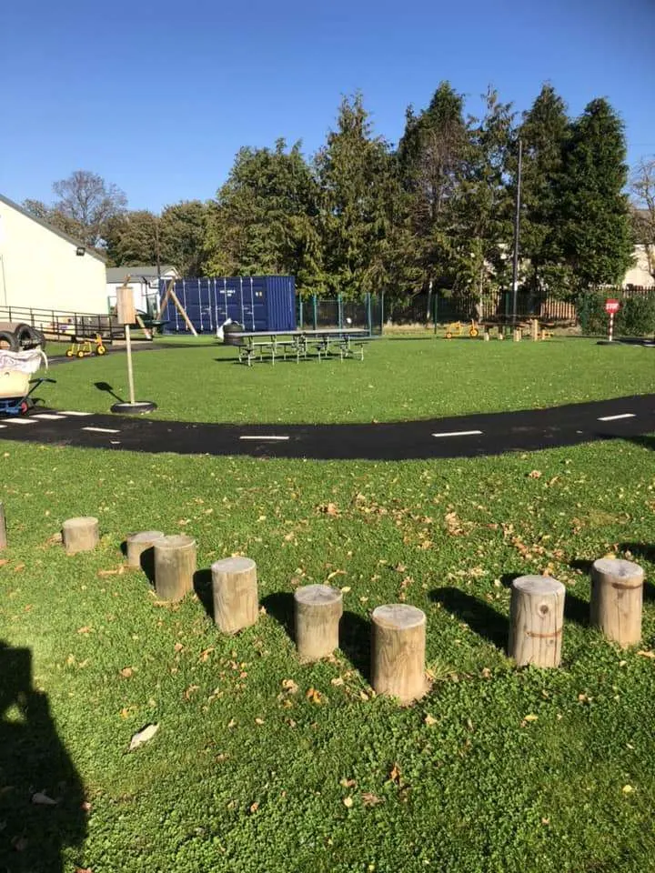 Outdoor play area with wooden log stepping stones on grass, trampoline, blue shipping container structure, and trees under clear sky.