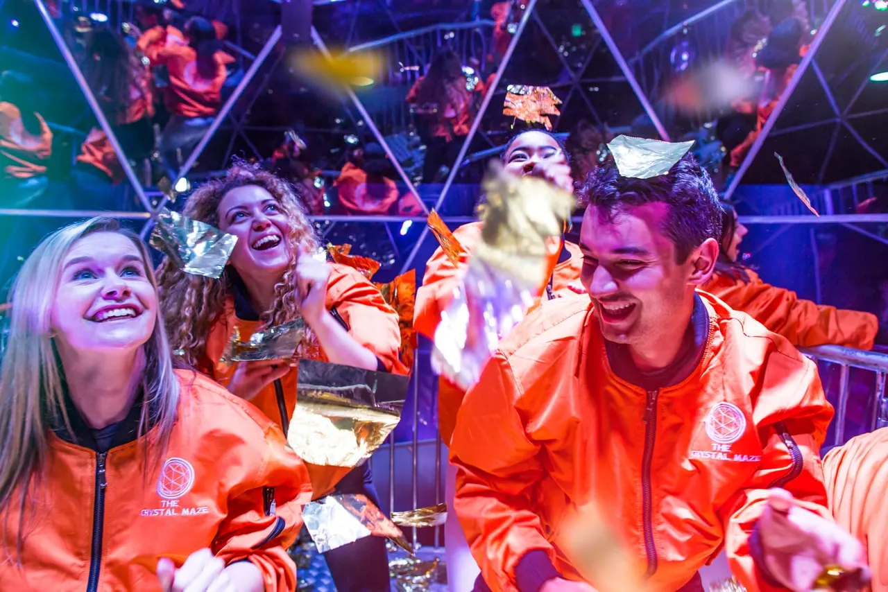 Group of smiling visitors in red jackets enjoying an interactive entertainment experience with colourful neon lighting and geometric set design.