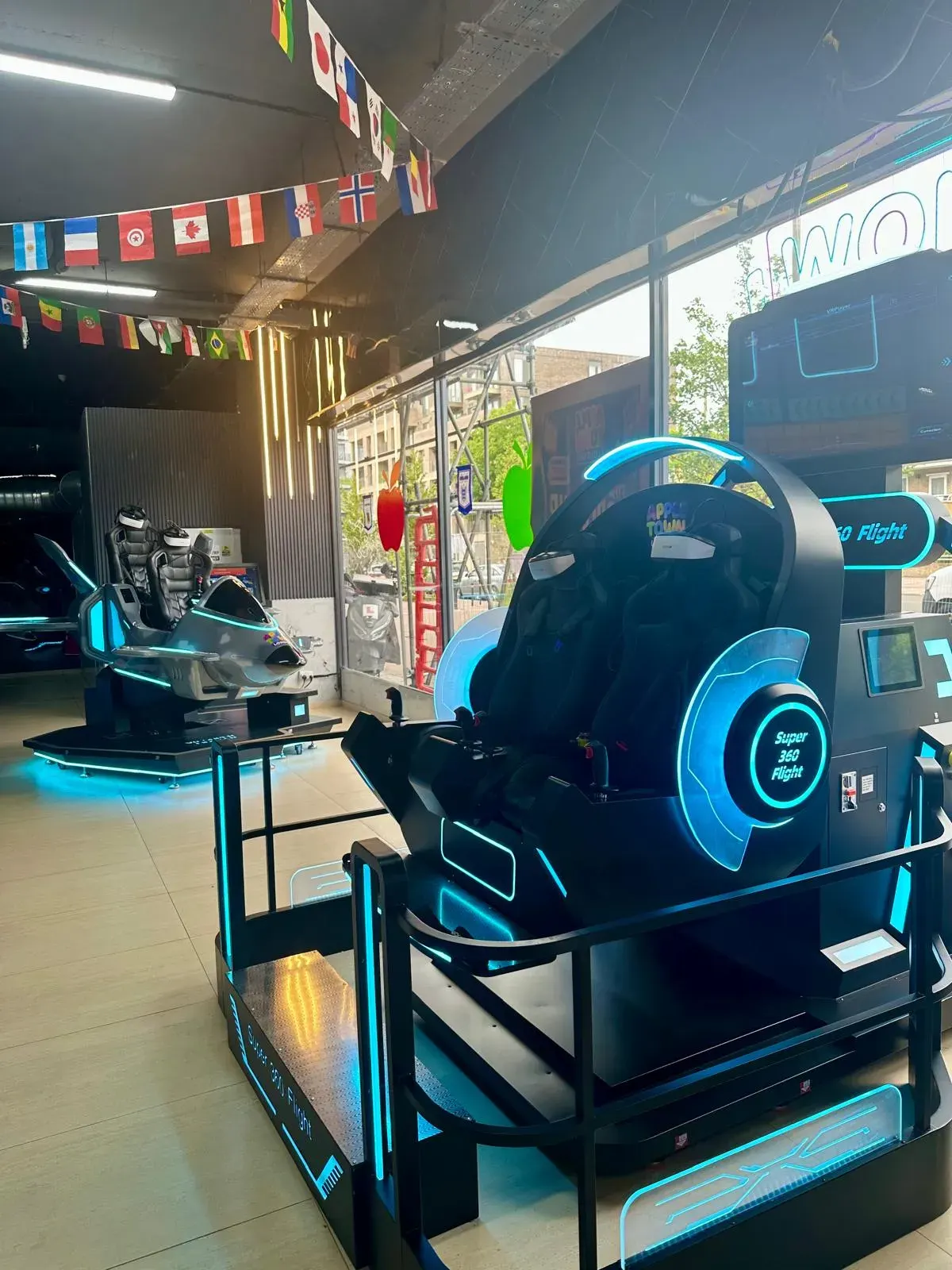 Virtual reality flight simulator arcade games with neon blue lighting in an indoor entertainment venue, international flags displayed overhead.
