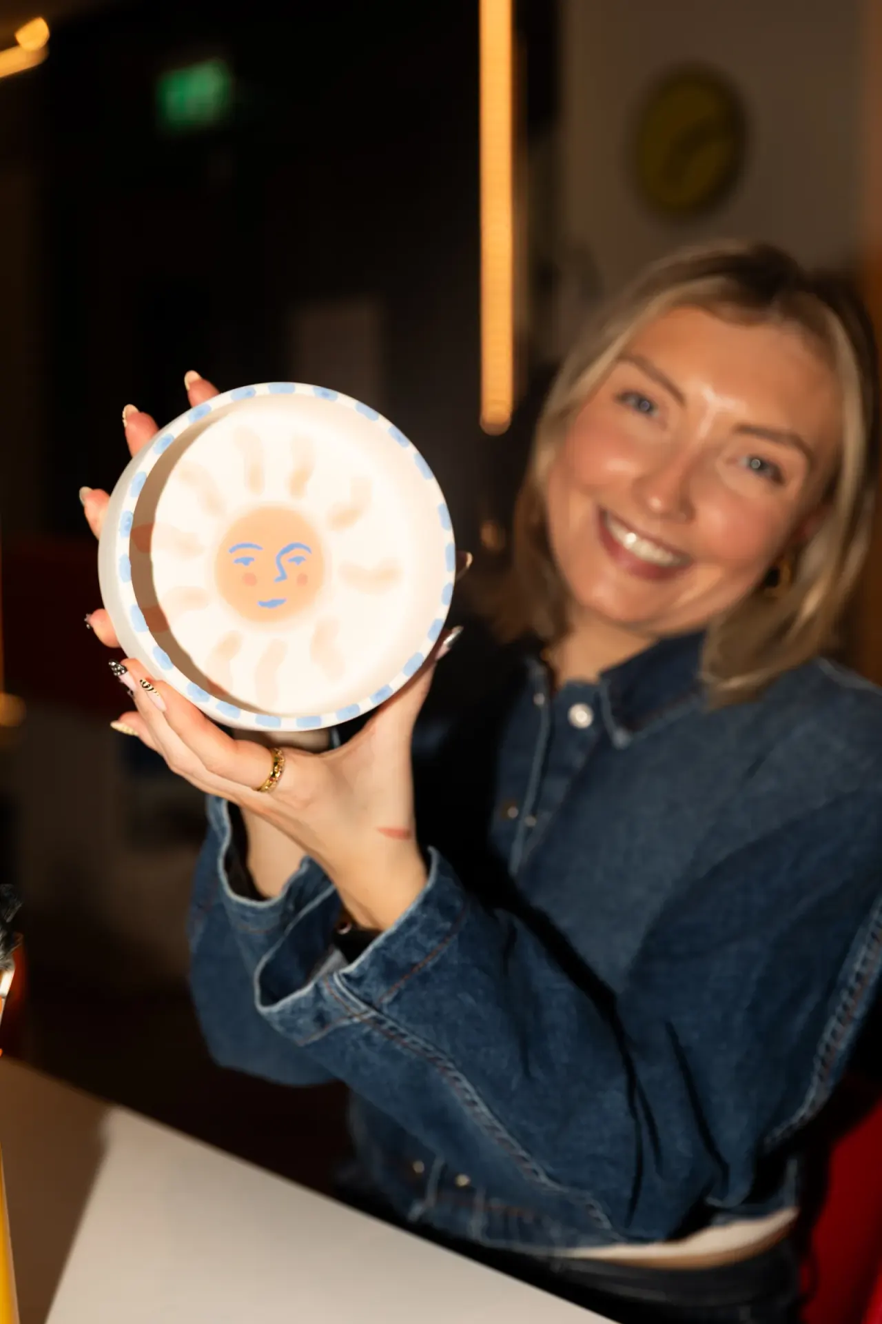 Woman holding a glowing decorative moon light with a smiling face design indoors.