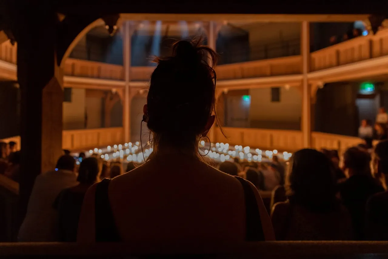 Woman seated in audience viewing a performance in an illuminated indoor theatre with wooden balconies.