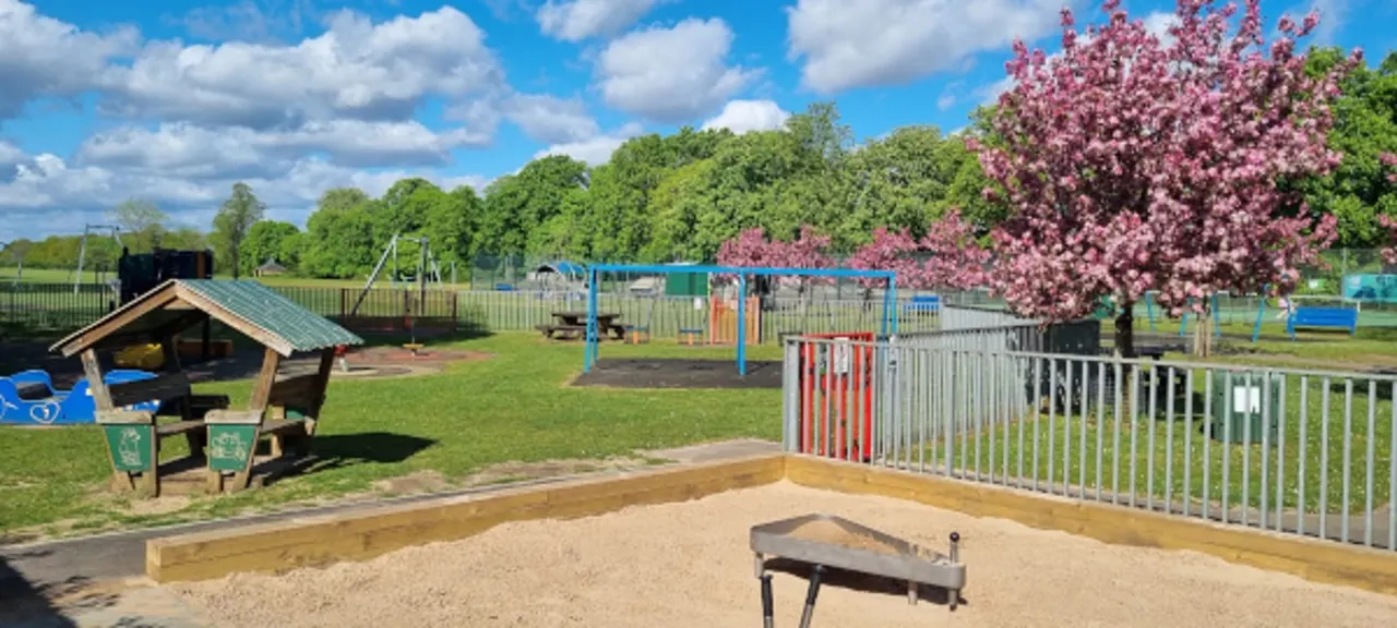 Sunny play area with sandy pit for games, picnic shelter, playground equipment and flowering trees under blue sky.