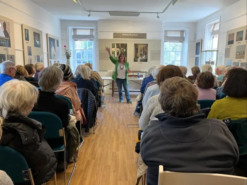 A presenter in a green top addresses a seated audience of mostly older adults in a bright, wood-floored hall with windows and display boards.