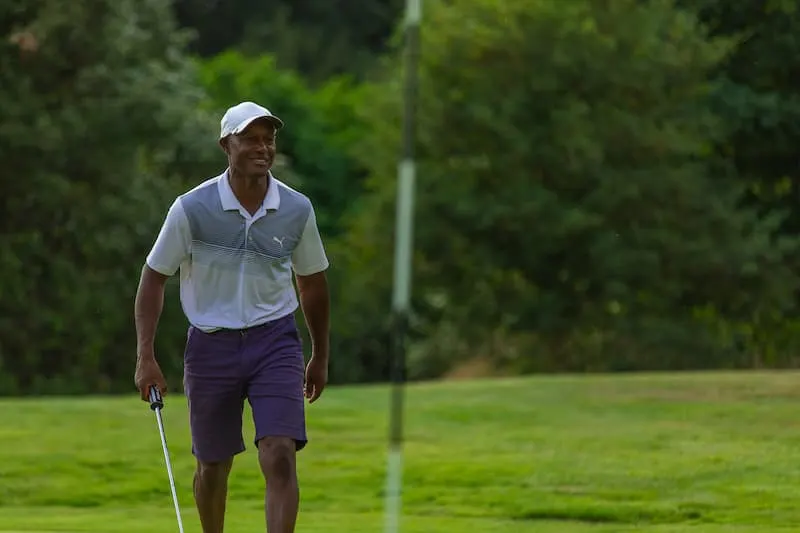 Golfer in white cap and polo shirt walking across green fairway with golf club.
