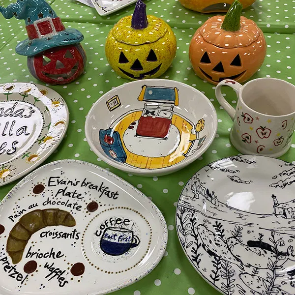 Decorated ceramic plates, bowls and mugs with hand-painted designs sit on a green polka dot surface alongside Halloween-themed ceramic pumpkin decorations.