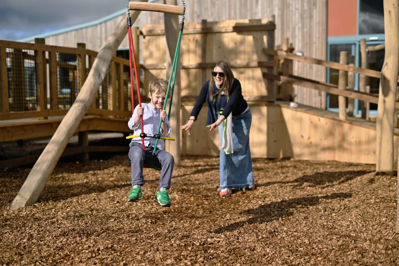A young child swings on a rope swing whilst an adult supervises in a wooden adventure playground.
