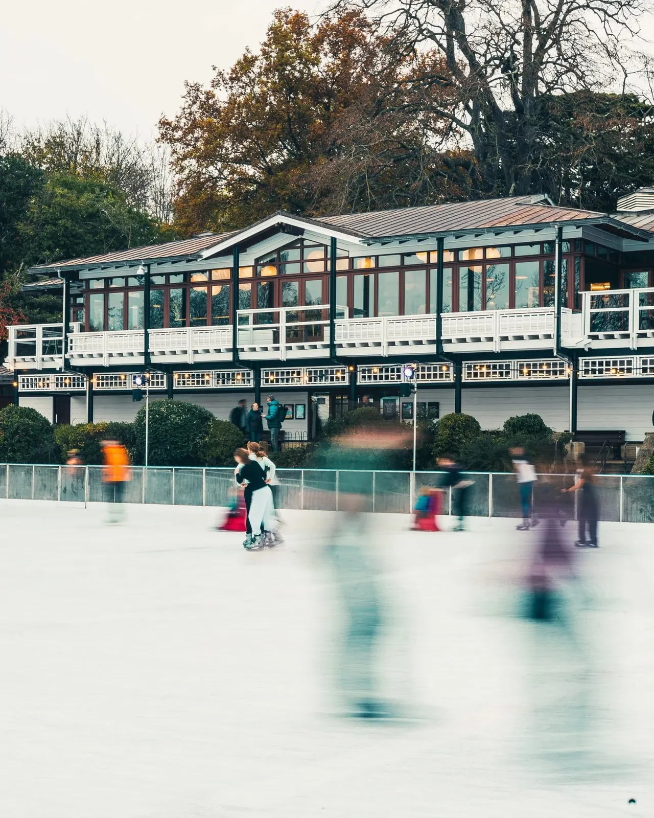 Multiple people ice skating outdoors in front of a modern pavilion building surrounded by autumn trees.