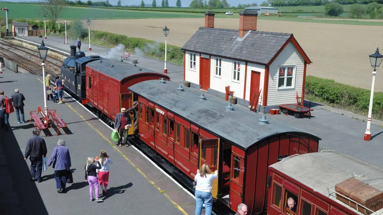 Visitors exploring a heritage railway station with a red steam locomotive, passenger carriages, and period buildings set in countryside.
