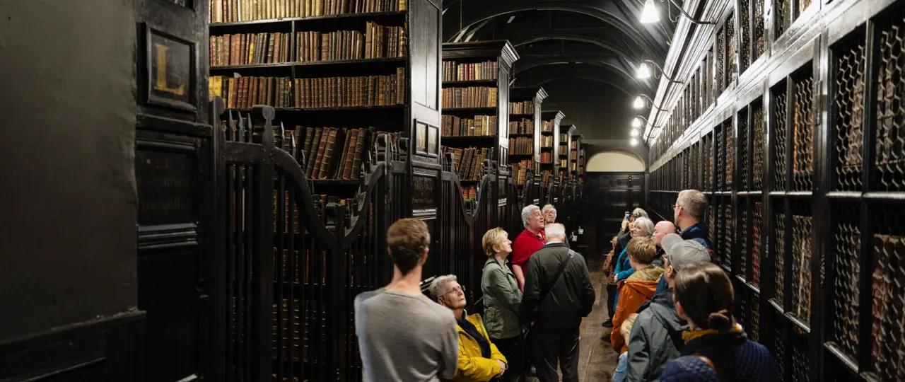 A group of visitors exploring a historic library corridor lined with old leather-bound books and ornate iron railings.