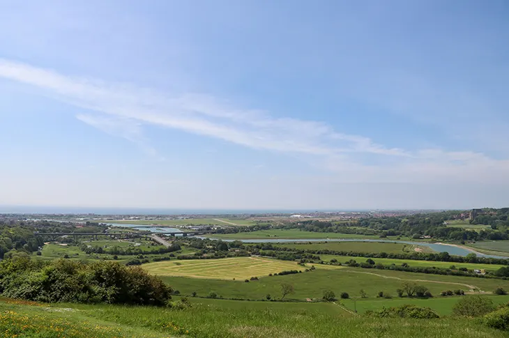 Elevated countryside view showing patchwork of green fields, trees and a winding river under blue sky.