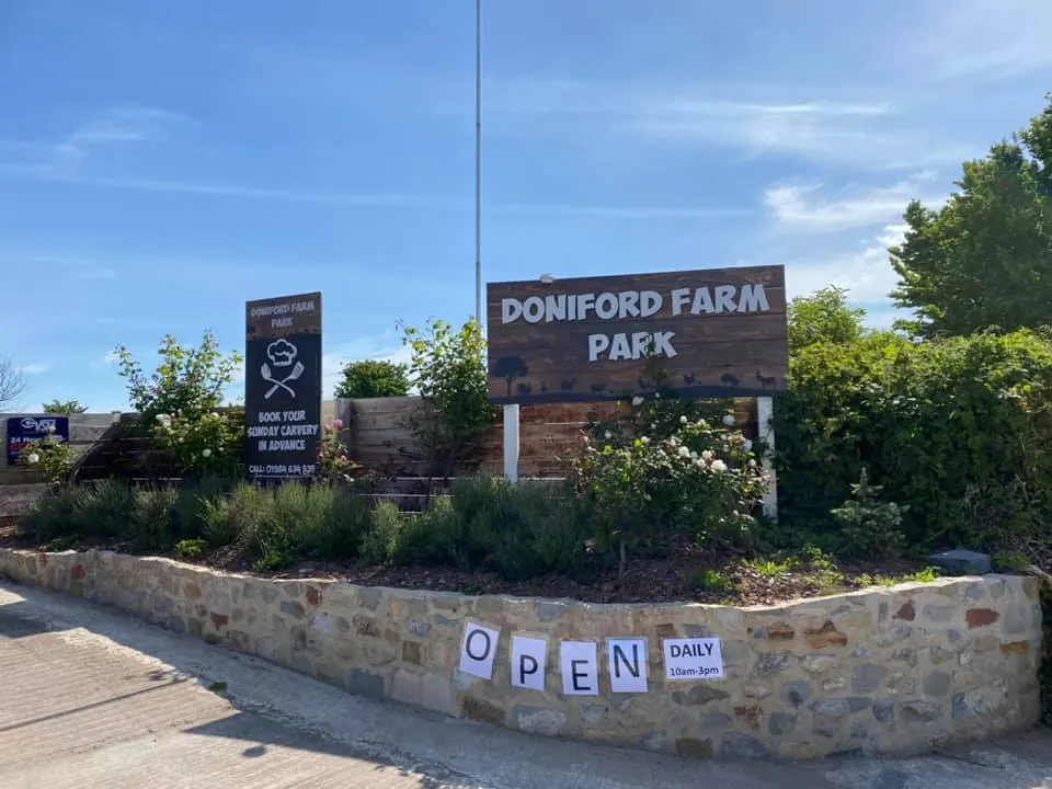 Entrance signage for Doniford Farm Park with wooden signs, stone walls, and landscaping under blue sky.