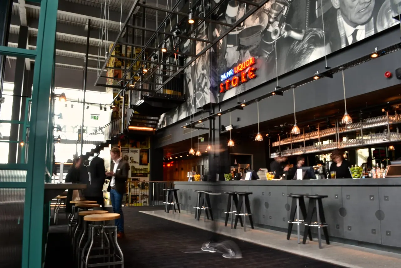 Modern bar interior with industrial ceiling, pendant lights, bar seating, and patrons in smart casual attire.