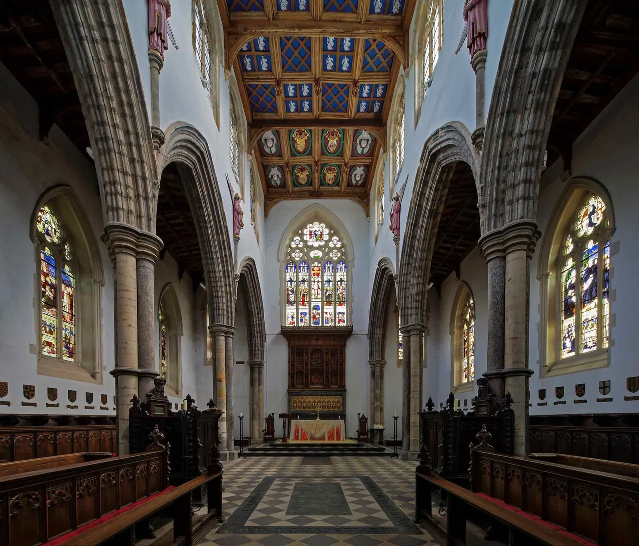 Interior of a medieval church with stone columns, wooden ceiling panels, stained glass windows and wooden pews lining the nave.