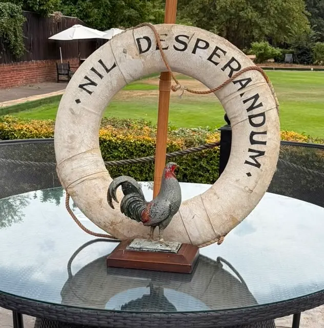 Decorative sculpture featuring a large stone ring with carved text and a rooster figurine, displayed on a table in a garden setting.