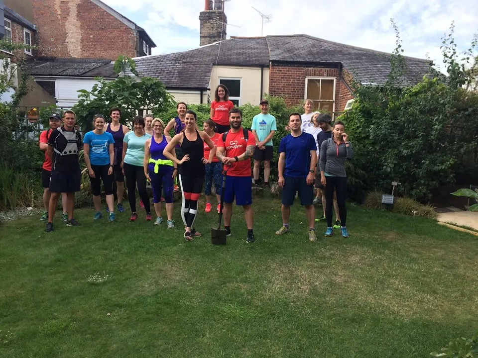 A group of about twenty people in athletic wear standing together on a lawn in front of a brick cottage, appearing to gather for a fitness or community activity.
