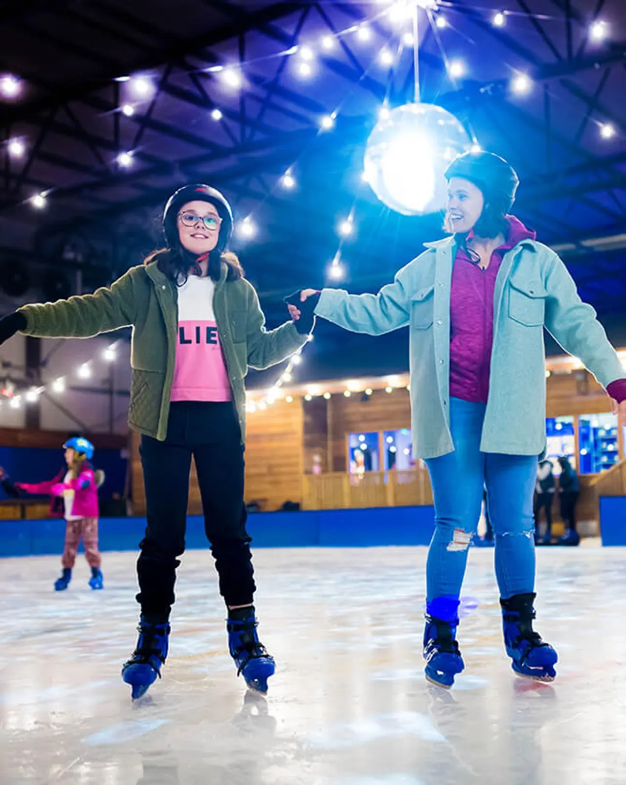 Two children ice-skating hand-in-hand on an indoor rink at night, surrounded by twinkling lights.