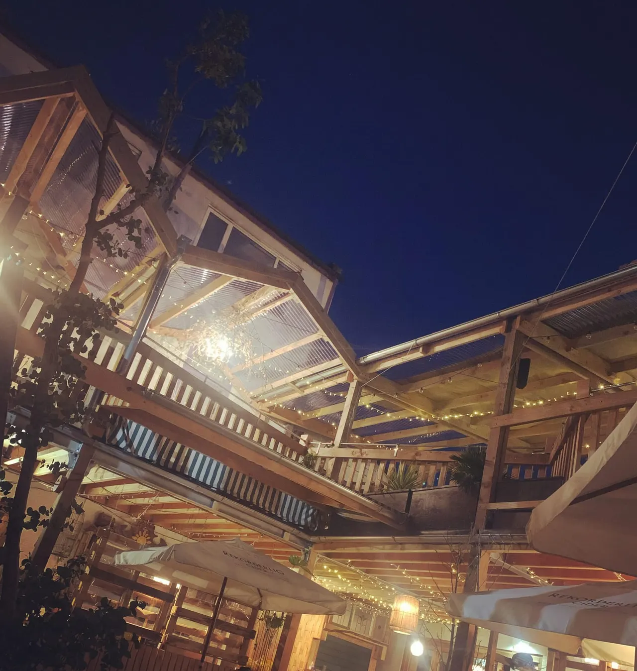 Illuminated wooden deck and pergola structures with string lights at dusk, overlooking an outdoor leisure venue.