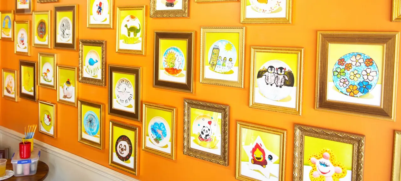 Gallery wall of children's artwork and paintings in various gold and brown frames hung on a bright orange wall.
