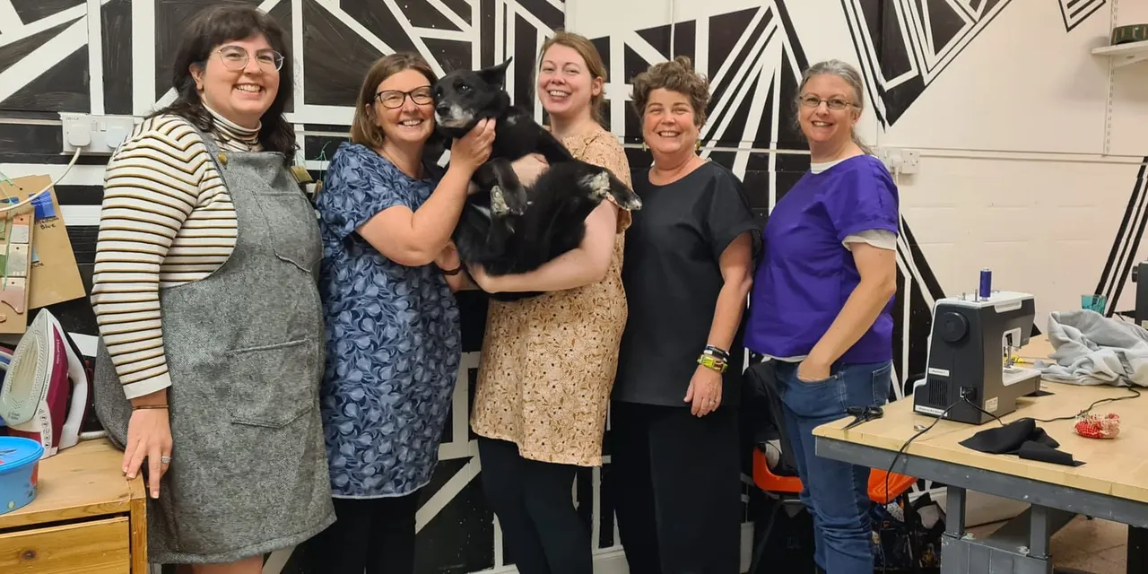 Five women posing together in an art studio, one holding a black dog, with geometric artwork on the wall behind them.