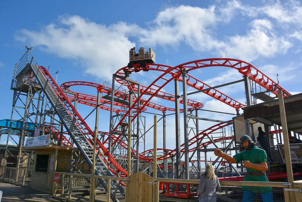 A roller coaster with red track and grey steel supports under blue sky, with visitors nearby at the amusement park.