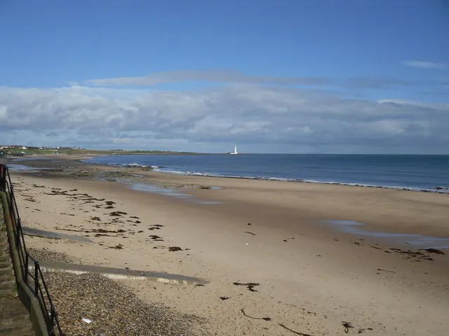 Wide sandy beach with shallow pools and exposed rocks at low tide, sailboat visible on horizon under cloudy sky.