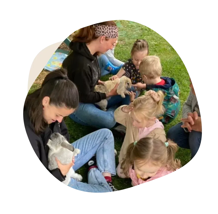 An adult and several young children sitting on grass, petting and handling small farm animals.